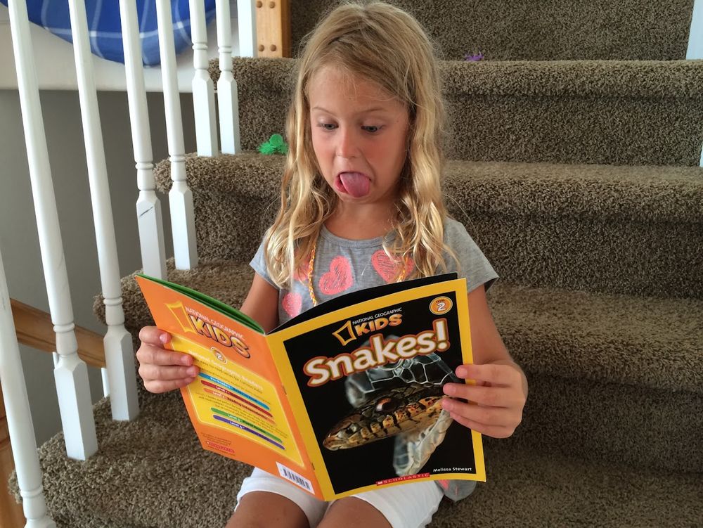 Our Daughter on a set of brown carpeted stairs with a Kids National Grographic magazine in hands. cover photo emblazoned with the headline "SNAKES!" and her response captured as thusly 😬