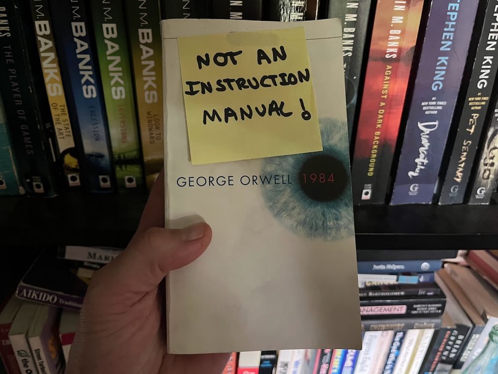 A copy of George Orwell's novel "1984' held in my left hand in front of a bookshelf with various other books including several Iain M. Banks novels; book is white with blue and red text, a close up view of an eye with black pupil and blue cornea, and a yellow sticky note attached with black handwritten marker text as follows" "NOT AN INSTRUCTION MANUAL!"