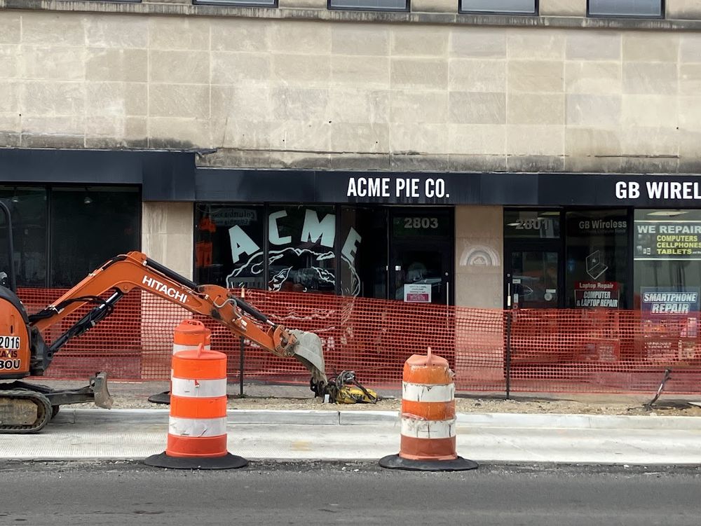 A construction fence and barrels along Columbia Pike in Arlington VA with a store front for "ACME PIE CO." in the background