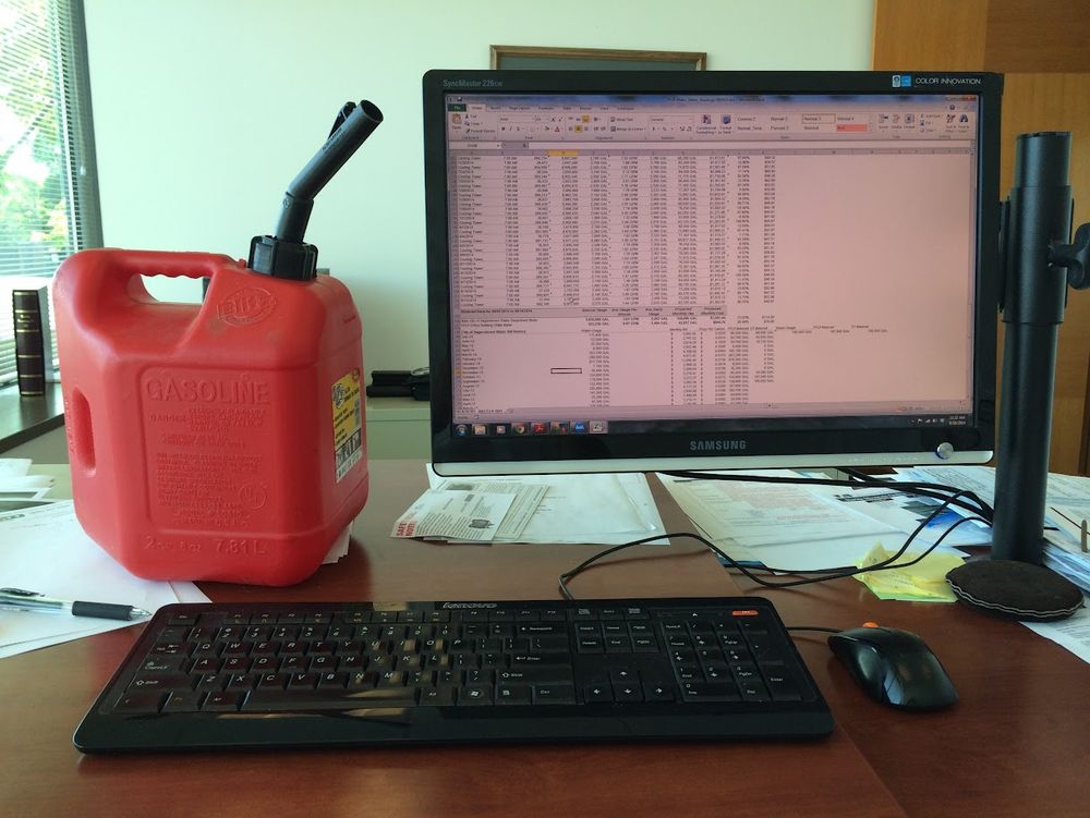 A red gas can on a brown desk, a black keyboard in front and a computer screen in the background with a spreadsheet open