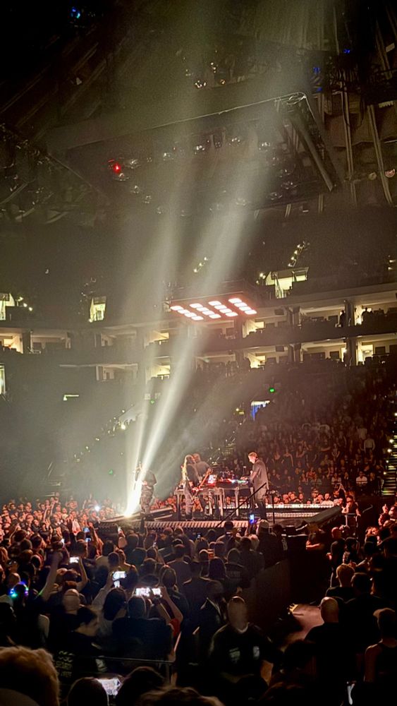 A picture of a crowd of people surrounding a small stage with four musicians playing on it bright lights shine up from the crowd onto the stage