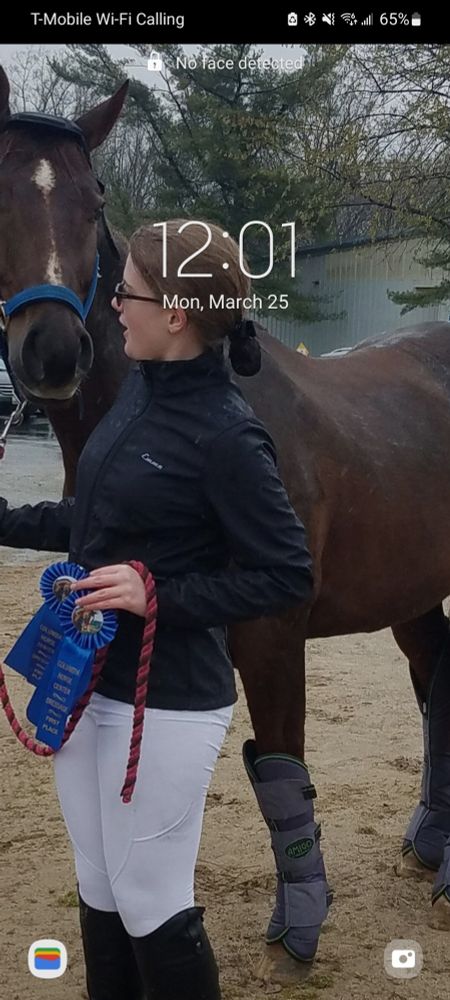 A woman with glasses holding two blue ribbons, and the lead rope of the chestnut horse behind her. She is turned to look at the horse.