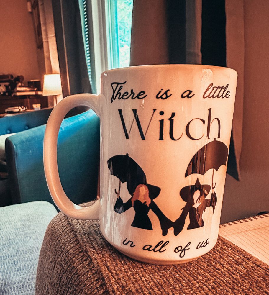 A coffee mug featuring a Practical Magic design sitting on a couch arm rest