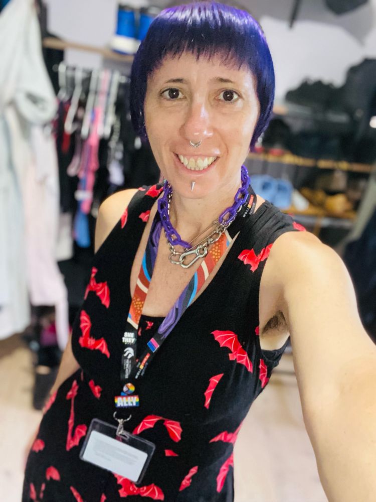 Image of Jackie from the hips up wearing a black dress with red bats. I also wear an indigenous lanyard with pride pins and a purple, chunky choker chain and a silver chunky choker chain.