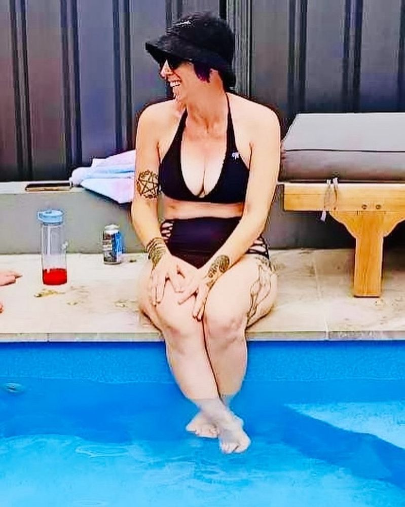 Another photo of me sitting on the edge of the pool with my feet dangling. In this time I had is turned to the side and I am laughing. I am wearing a black bikini, a black bucket, hat and black sunglasses.