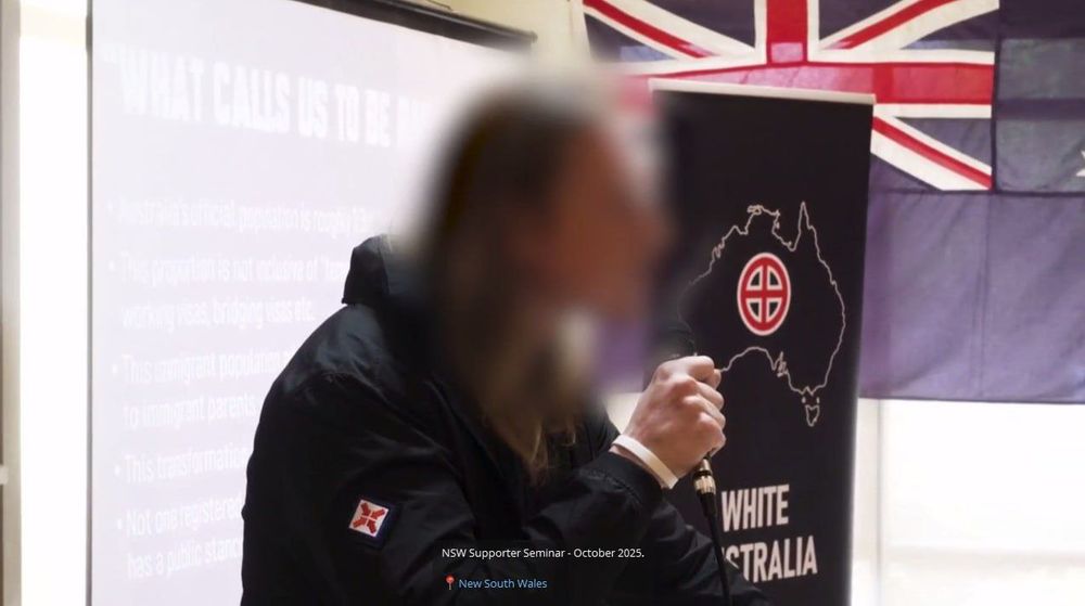 Oscar Tuckfield blurred but recognisable in a patched NSN jacket, giving a speech to NSN/White Australia's supporters at their recent NSW seminar night in October. The powerpoint slide behind him has anti-immigrant talking points and he's holding a microphone.