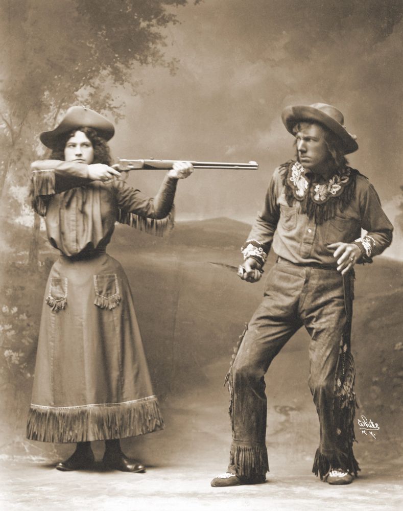 historical photo of a woman in fringed western attire pointing a gun at a man in costume (promotional photo of Annie Oakley)