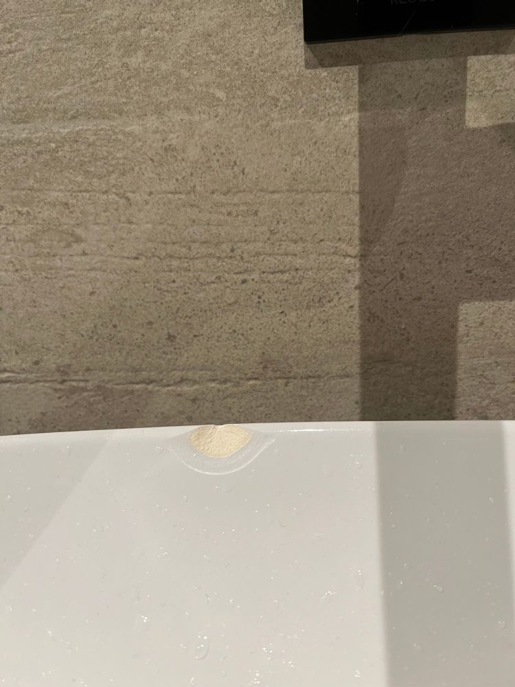 Abgeplatzte Stelle  am Rand eines weißen Porzellanwaschbeckens. Im oberen Teil des Bildes ist eine betonartige beige Fliese zu sehen.