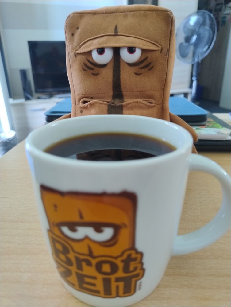 Ein Bernd das Brot Plüschtier sitzt hinter einer großen weißen Tasse, auf welcher comicartig das Konterfei des Brotes zu sehen ist, sowie der Schriftzug BrotZEIT. Der Inhalt der Tasse ist dunkelbraun bis schwarz.  Bernd spiegelt sich etwas auf der Oberfläche der Flüssigkeit der Tasse (es ist Kaffee).