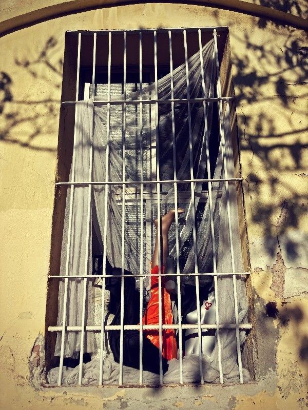 A mannequin's leg in orange trousers appears upwards behind a barred window with sheer white curtains, next to a white stuffed animal, in a weathered Barcelona building… #SilentSunday © 20251109 Gilles Denizot