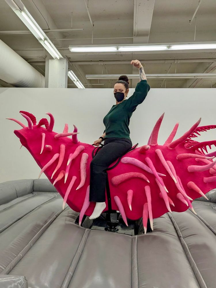A woman with a mask rides a bright pink mechanical sea slug sculpture