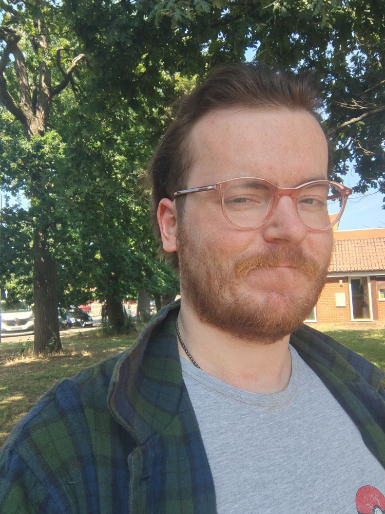 An image of me with shorter hair, I'm wearing a grey top and have a green and blue checked overshirt over the grey top. there are trees and a building behind me and the sky is blue.