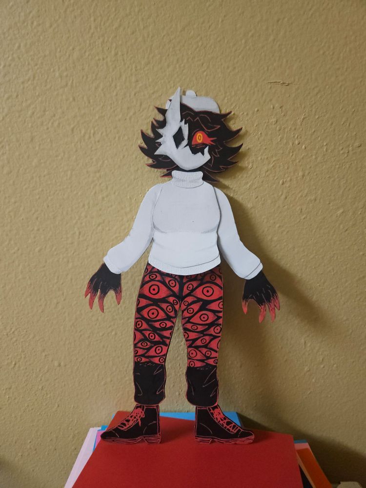 A detailed paper doll of Folly, an antagonist from the game Regretevator. She is a humanoid character who is solid black in color, with medium length, spiky hair, clawed fingers with red tips, and a single visible eye that is bright red with sharp lower lashes. The iris and pupil of her eye are made up of two bright yellow concentric rings.

She wears a white turtleneck sweater, a white beret on her head, black pants with realistic red eyes all over them, and black boots. On her face she wears a broken mask that resembles some sort of animal. The mask has a single, diamond shaped hole for an eye, three spikes on the outer edge that look like fur, and a single, tall ear pointing upwards. The missing piece of the mask reveals her eye.

The doll is standing on a stack of papers and is leaned against a tan wall, to show the contrast better than the other images.