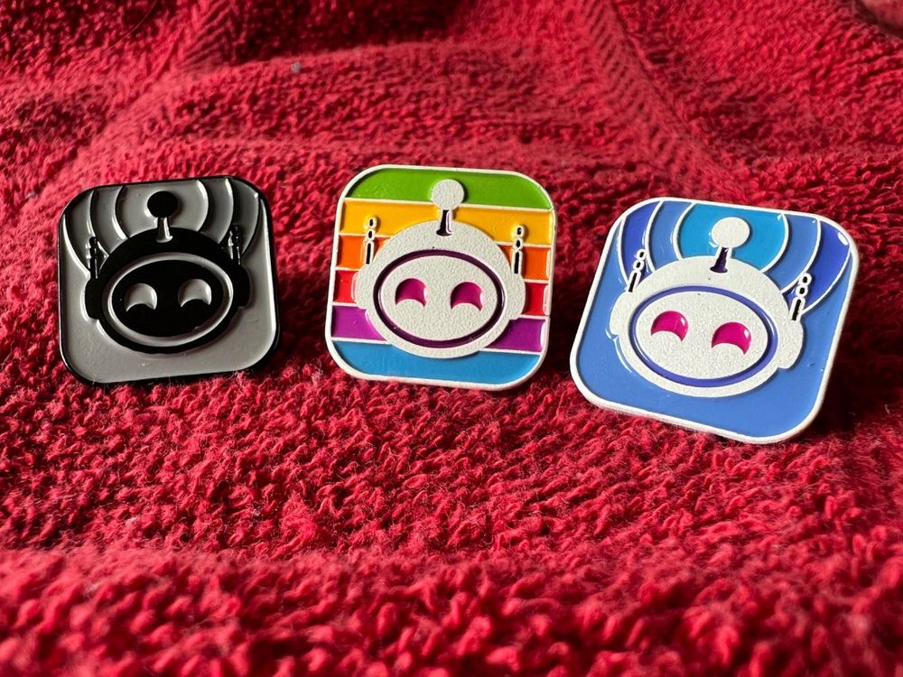 Three enamel Apollo pins on a red blanket background, the pins are dark themed, rainbow, and normal blue and about 1 inch in size