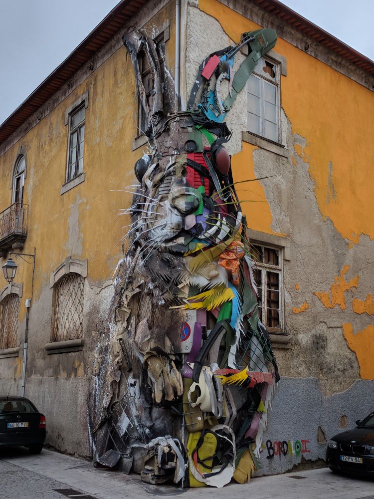 Un coin de rue à Porto. L'angle d'une maison d'un étage a été peint et recouvert d'un certain nombre de déchets plastiques colorés pour former un gigantesque lapin assis, très réaliste.
Oeuvre d'Artur Bordalo, alias Bordalo II, dont "l'œuvre se compose principalement de grandes installations et de fresques murales réalisées à partir de déchets recyclés, visant à mettre en lumière le gaspillage et la surconsommation dans notre monde actuel" (wikipédia).