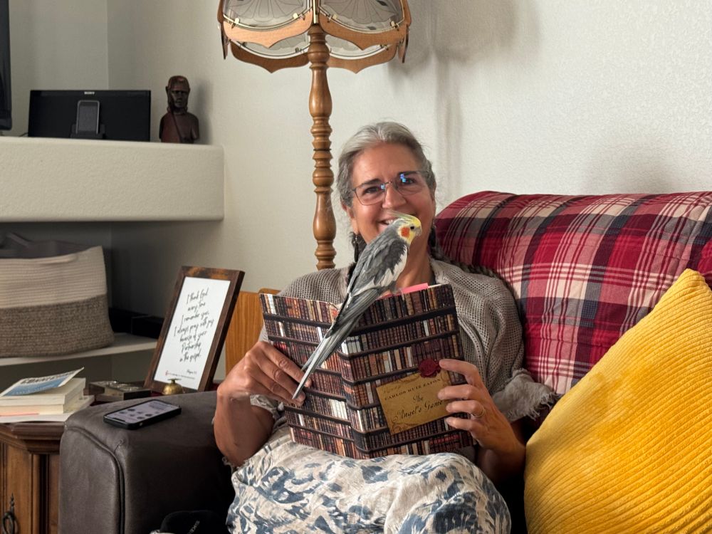 I am sitting on a couch reading a book that a cockatiel is perched upon.