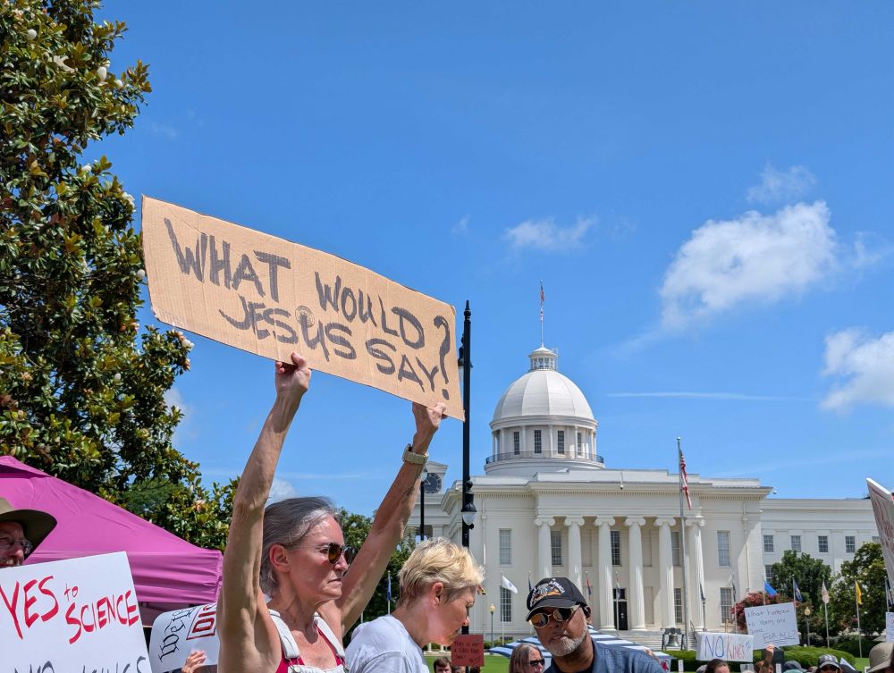A woman holds a sign in front of the Alabama State Capitol saying “What Would Jesus Say?”