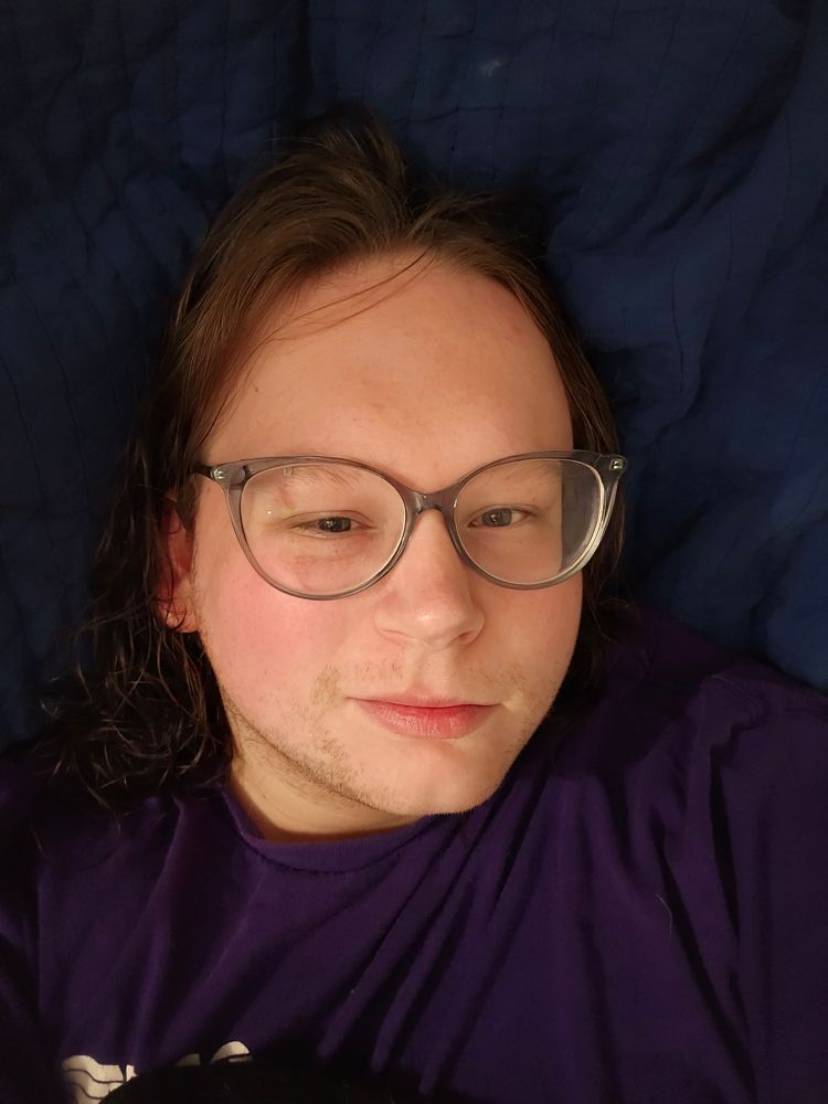 Sophi laying under a blanket with her glassss on, wet and wavy hair drape down her neck. She is wearing a purple shirt and is smirking. Slight stubble is visible