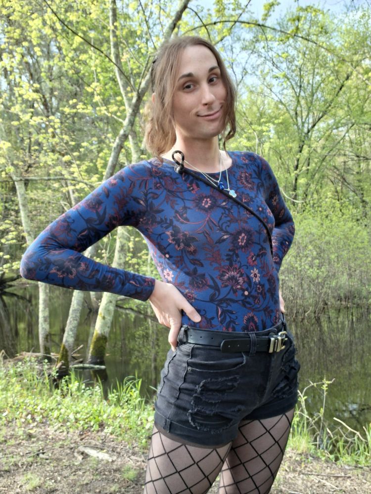 Picture of Cassie from thighs up standing with her hands on her hips in a very green forested area
She's wearing a dark blue and floral print, body conforming top, black distressed jean shorts and diamond pattern black tights, she's wryly smiling