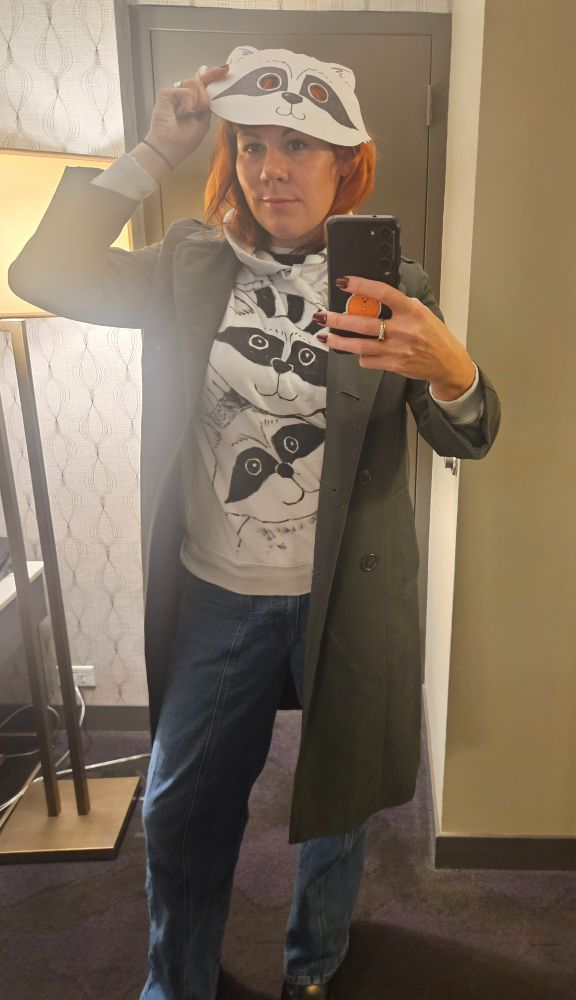Me in a white hoodie with two raccoons on it with me and a raccoon mask and green trench coat