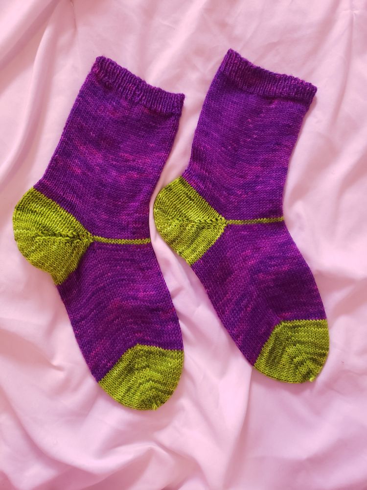 A pair of knitted socks laid flat on a white blanket. The body color is purple and the heels and toes are in a bright green.