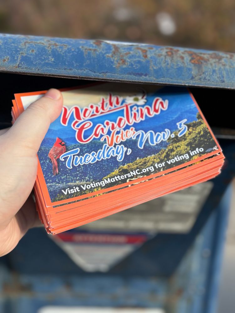 A hand putting a stack of 200 postcards into a mailbox.  The postcards say North Carolina Votes Tuesday Nov 5 visit votingmattersnc.org for voting info on them. 