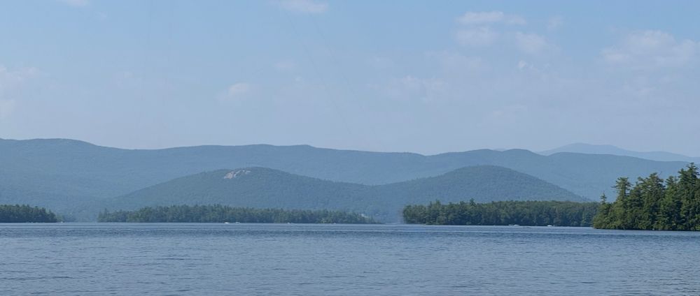 A lake in the foreground, leading out to some mountains in the background. Of of the lower mountains has a rock face, while rest are covered in trees. Squam Lake, New Hampshire. 