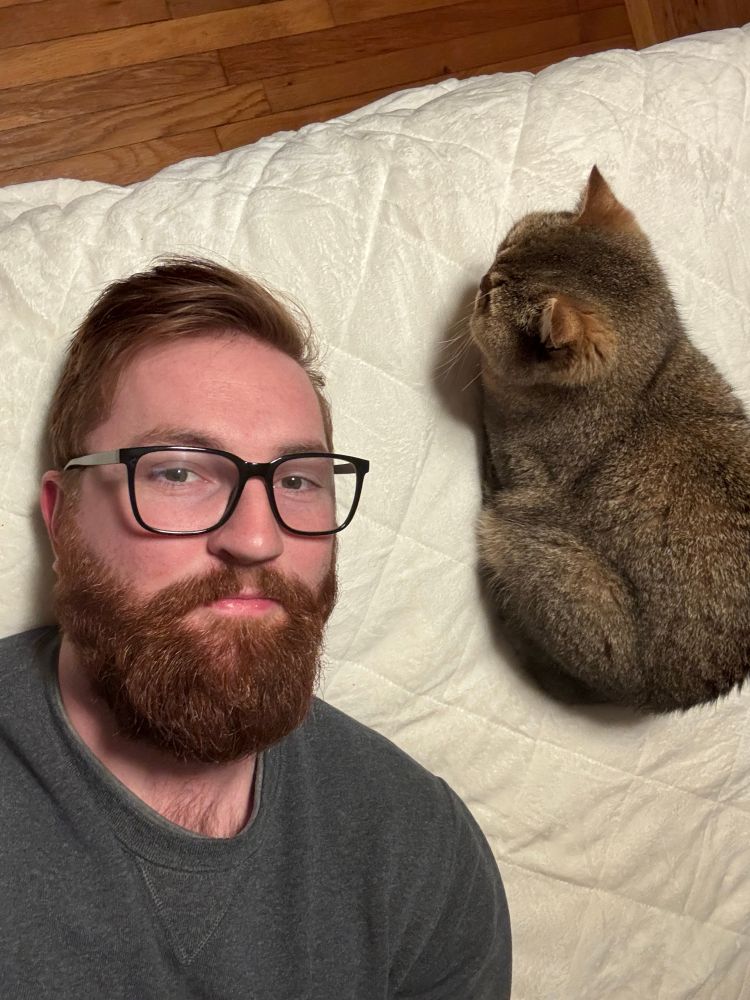 Me, a white man with red hair and a beard, wearing thick rimmed black glasses and a grey sweater, laying on a white blanket next to Wally, a brown british shorthair cat
