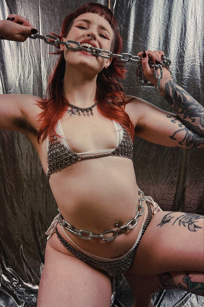 Stevie wearing a chainmaille bikini with a white bikini under it, a chain belt made of heavy hardware store chain, Stevie is biting a separate link of chain while squatting 