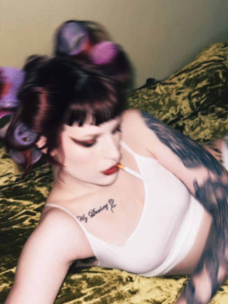 Stevie in a white tank top and rollers in their hair with a cig hanging out of their mouth laying on a bed kinda blurry 