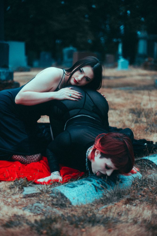 Stevie and Effy in a graveyard, Stevie is kneeling on all fours licking a grave stone while Effy rests their hands and head on Stevie’s butt looking seductively at the camera as if we’re trying to seduce you 