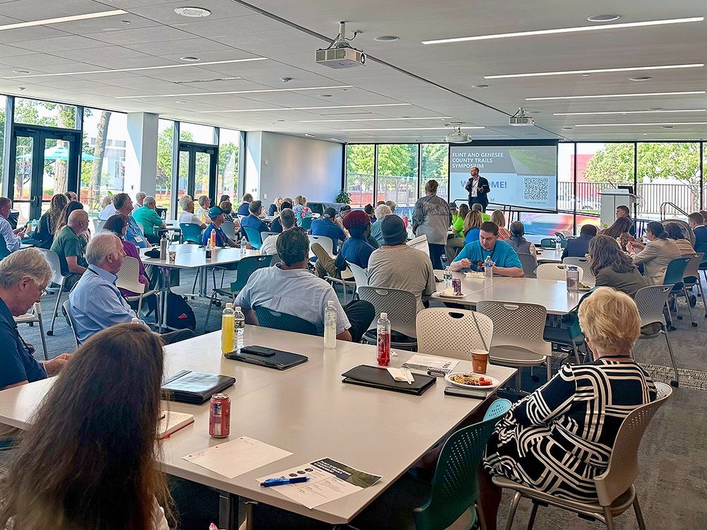 A large group seated at tables in a room with lots of natural light listen to a speaker presenting in front of a welcome slide with the text "Flint and Genesee County Trails Symposium."