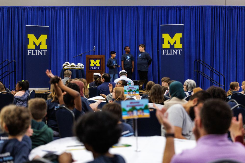Three boys stand on a stage with UM-Flint banners, talking to a room filled with children seated at tables.