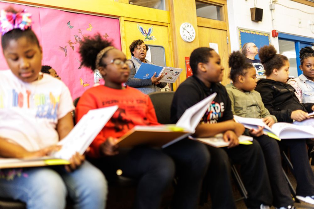 A row of kids sit in a classroom setting with open books in their laps, while a woman in the background reads aloud.