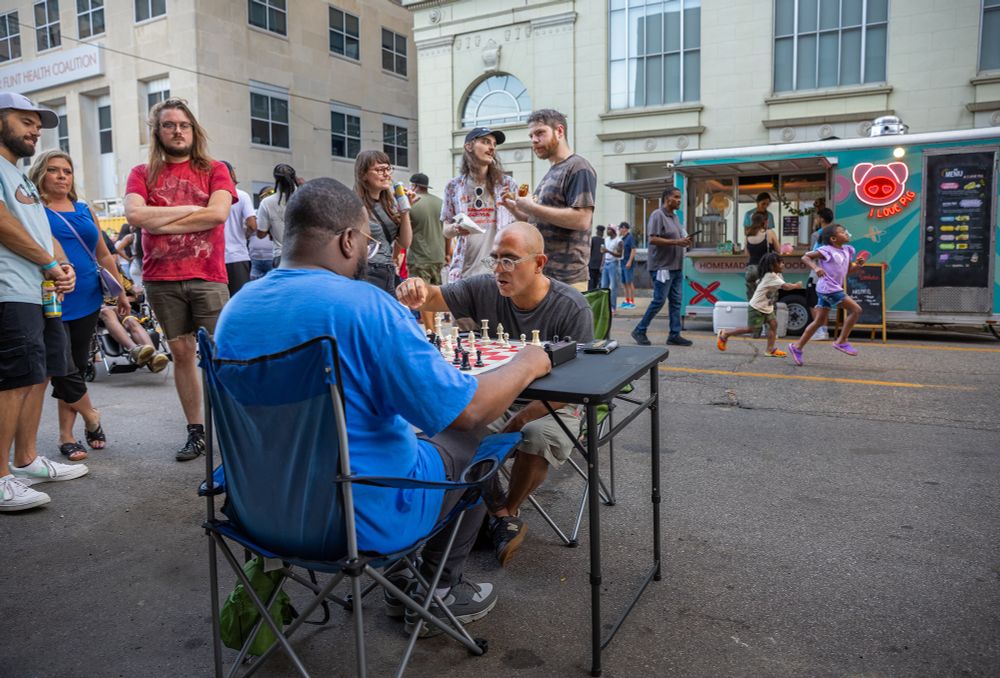 Two men play chess at a folding table in an outdoor alley as bystanders watch.