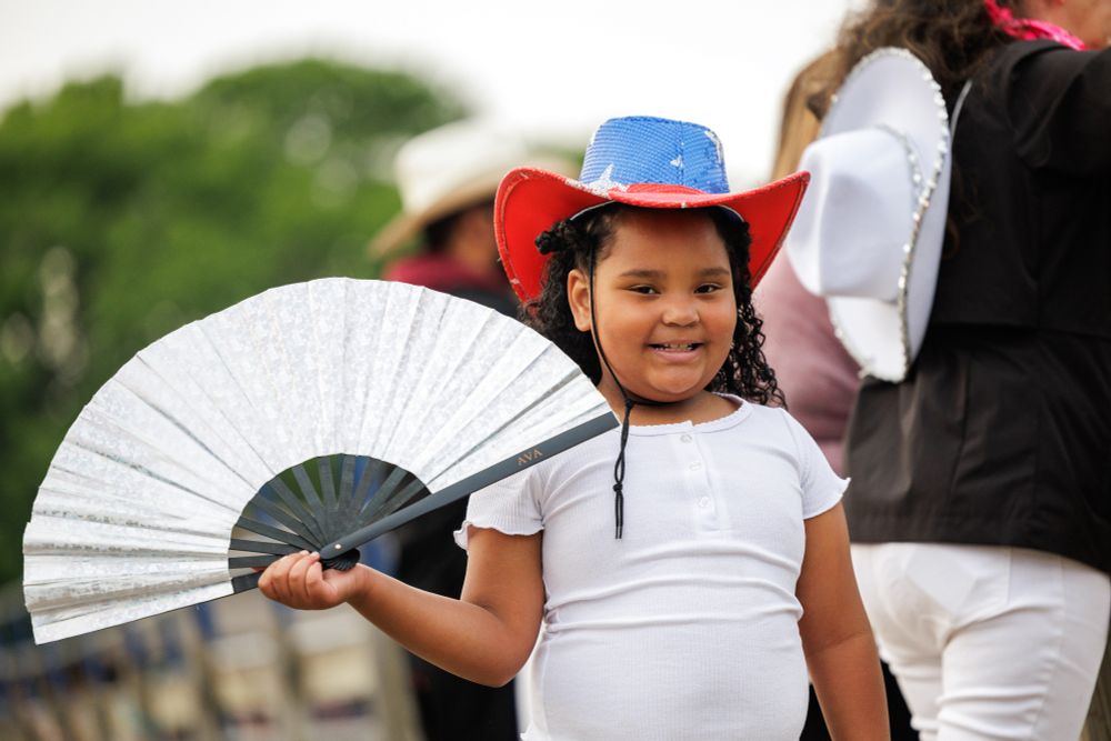 A girl wearing a red, white and blue sequined hat smiles as she holds up a large white fan.