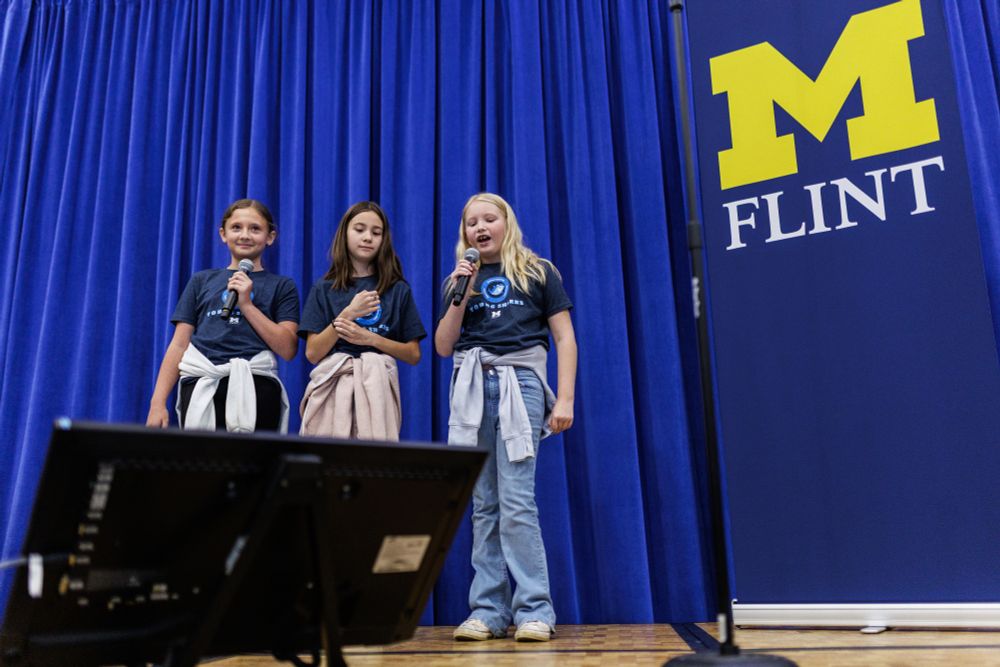 Three girls address an audience while standing on a stage with a UM-Flint banner.