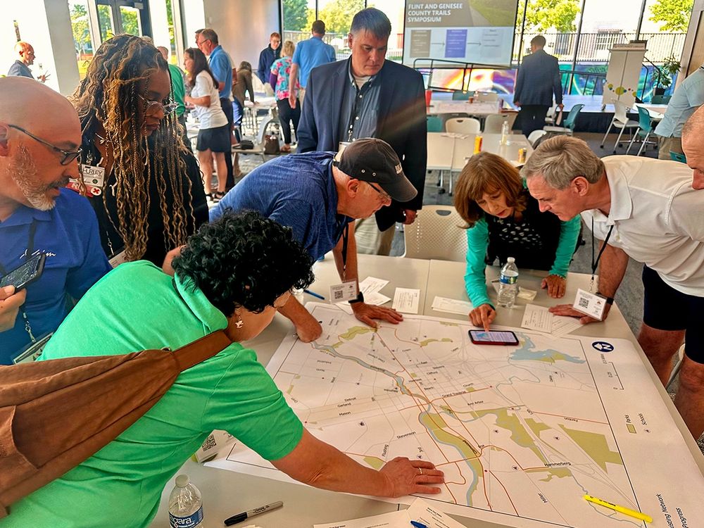 A group of people have a discussion over a large printed map spread out on a table. 