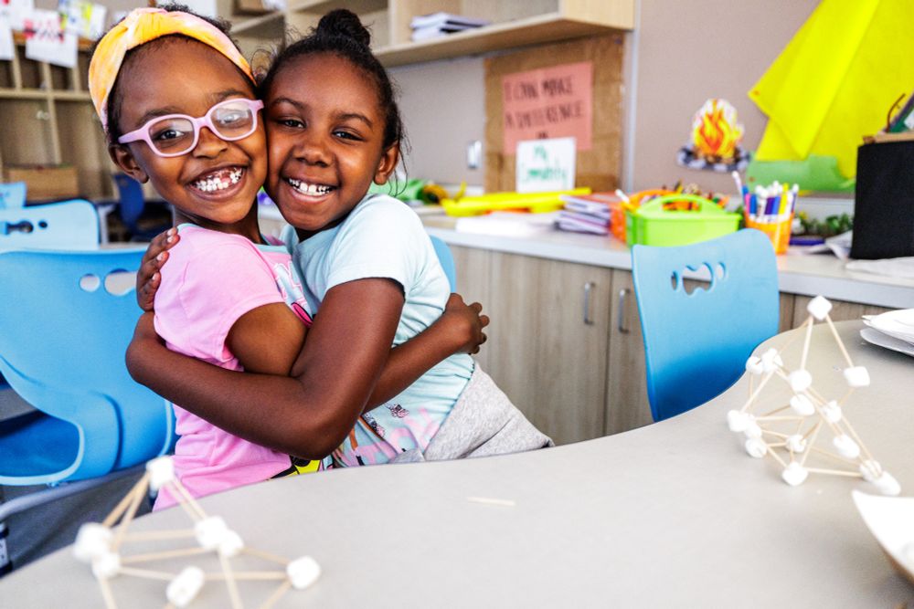 Two smiling girls take a break to hug while working on a marshmallow and toothpick STEM project.