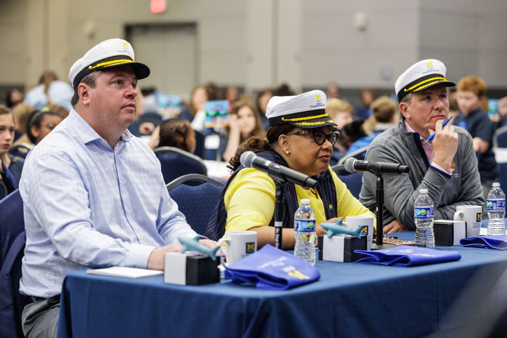 A woman and two men, all wearing captain hats, listen intently to presenters.