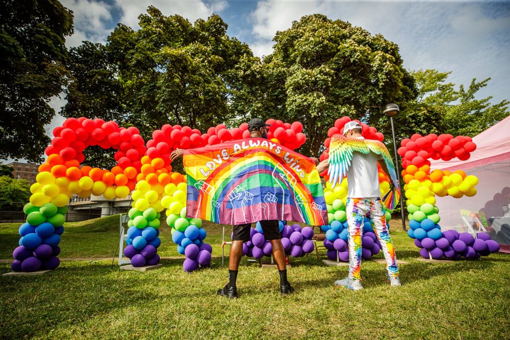 Two people in rainbow gear with their backs to the camera stand in front of a rainbow balloon sculpture that spells out pride. One person holds a flag that says love always wins, and one has rainbow wings.