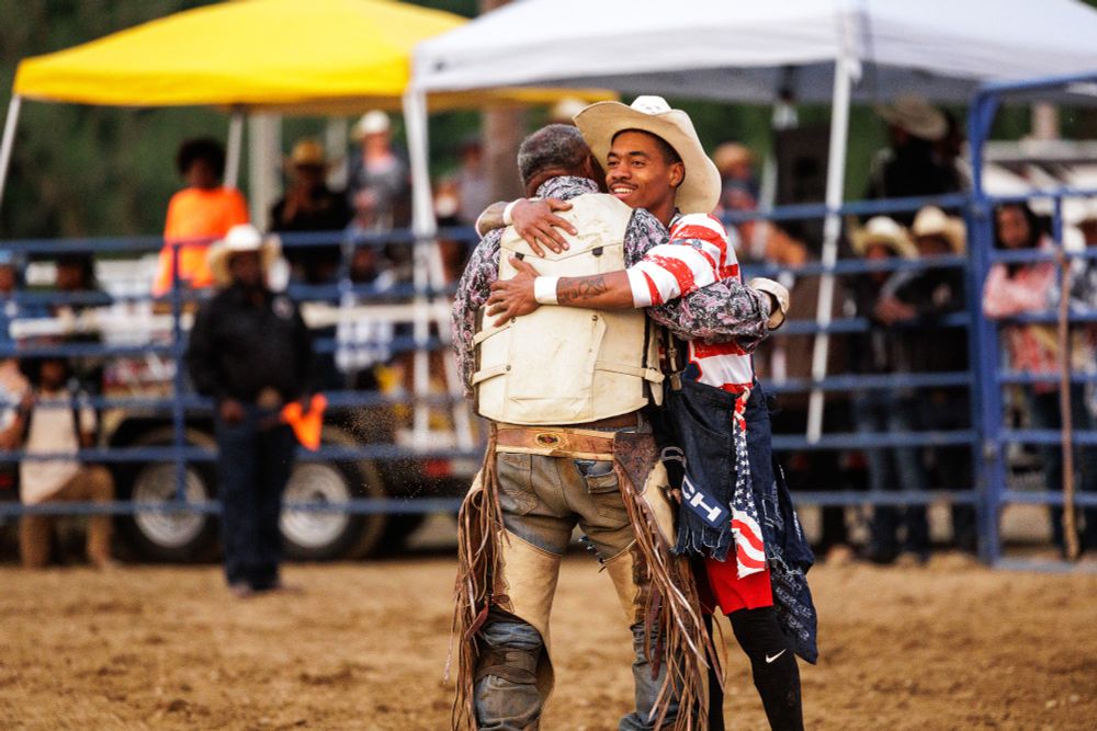Two men wearing rodeo gear hug while standing in fenced in fairgrounds.
