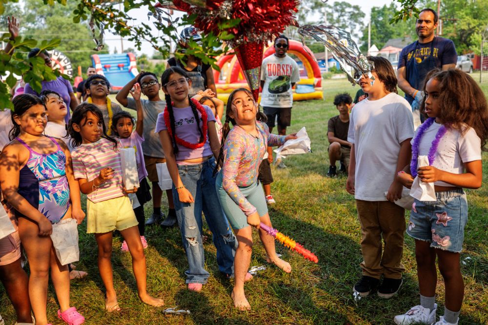 A girl looks up with determination at a glittering red and silver pinata as she prepares to strike it with a colorful stick. A group of children and several adults watch in the background.