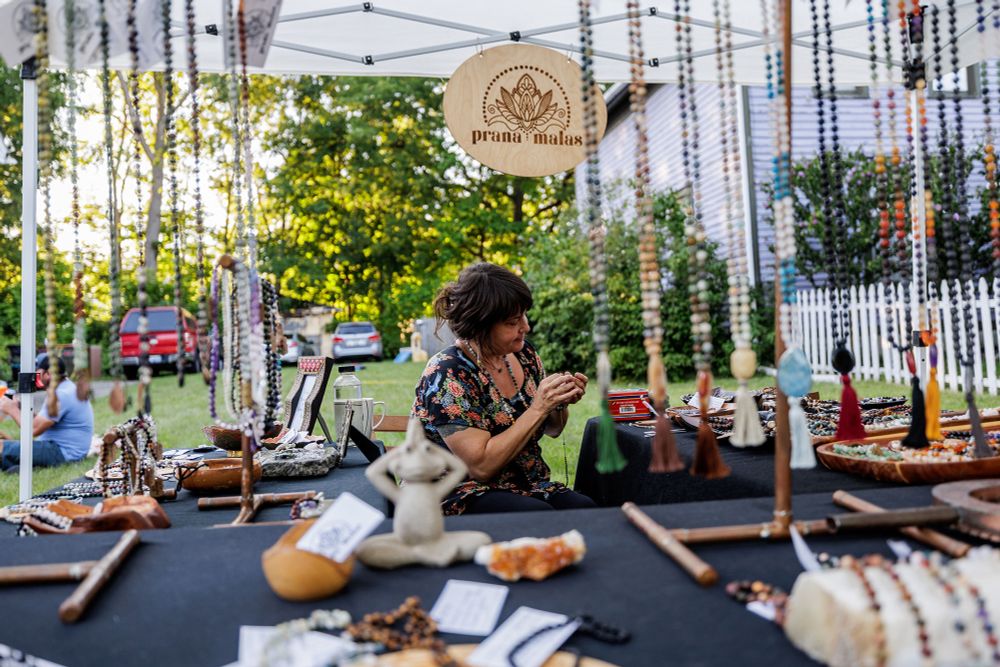 A woman works at a vendor booth under a sign that says prana malas. Beaded and decorative items are on tables around her and hanging from her tent. 