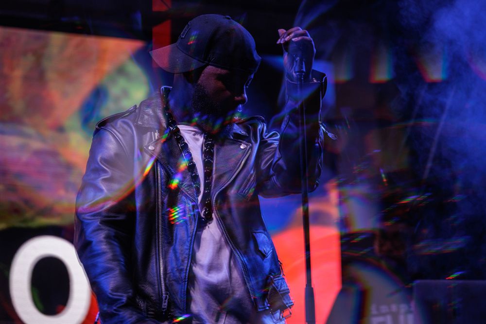 A man looks down and away as he stands in front of a microphone on an artistically lit stage.