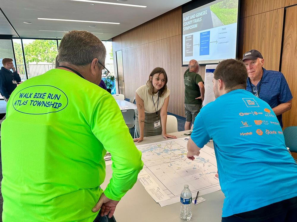 A group of people have a discussion over a large printed map spread out on a table. 