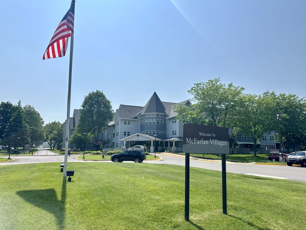 The exterior of McFarlan Villages building on a sunny day. An American flag and a building sign that says "Welcome to McFarlan Villages" are in the foreground.