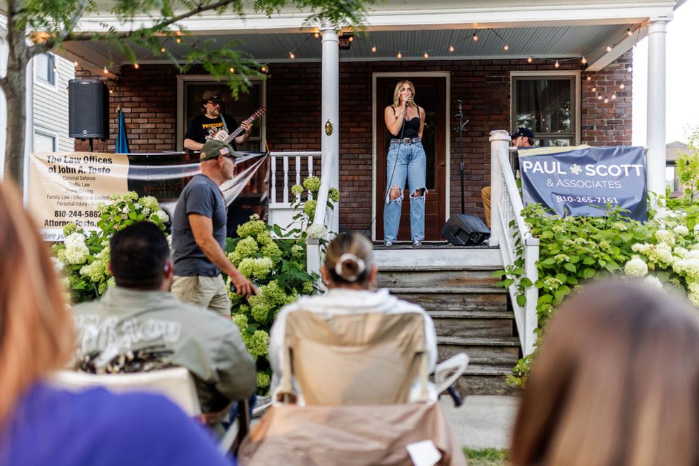 Performers use a porch as a makeshift stage while a seated audience watches.