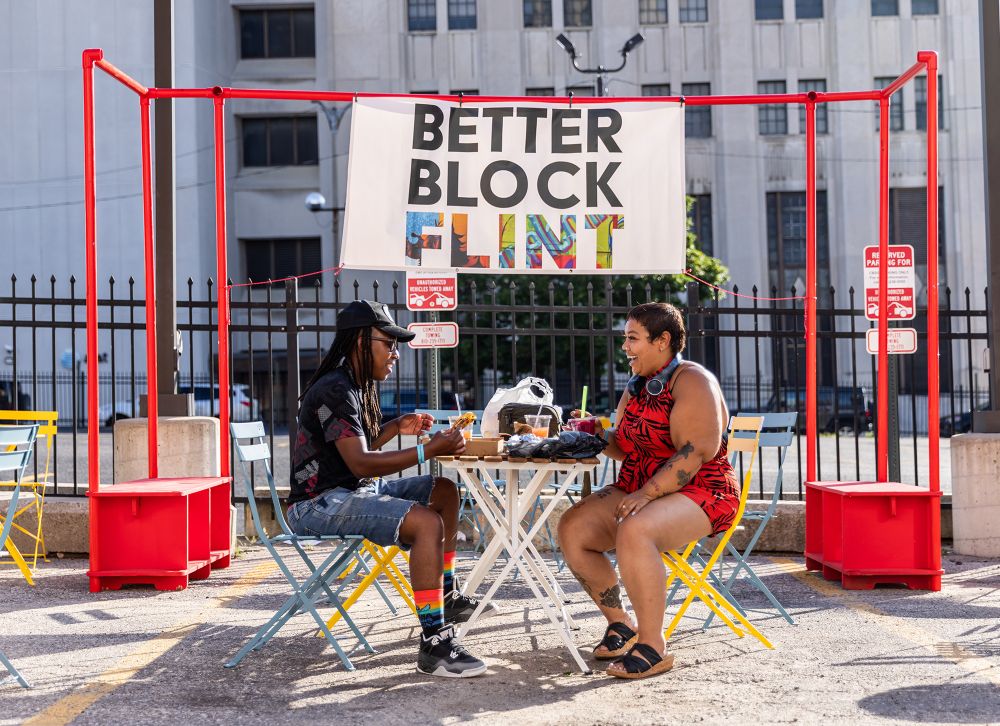 Two people eat and chat while sitting at a table in a colorful seating area in front of a "Better Block Flint" sign.
