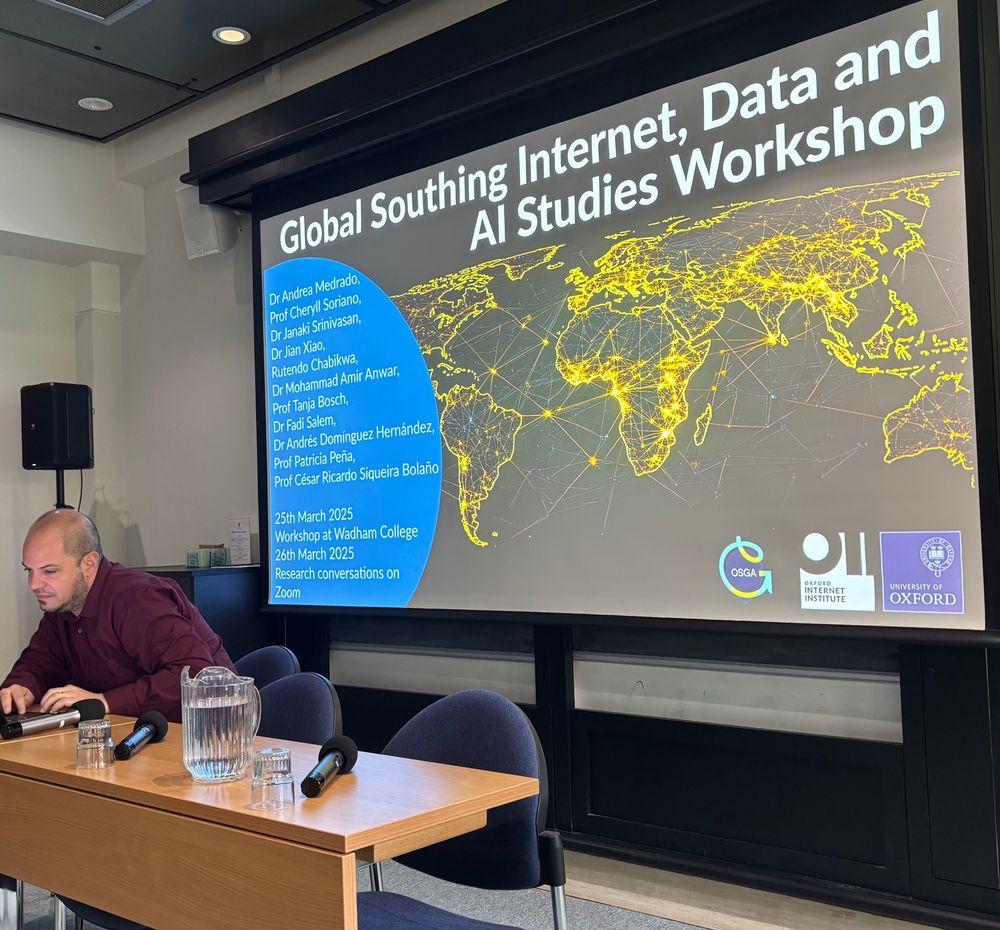 Close up photo of seminar room - Jonas Valente sits at a table in front of the projector screen. The table has microphones and a jug of water. Jonas looks at his laptop. on the projector screen behind him, the title screen of the workshop reads 'Global Southing Internet, Data, and AI Studies Workshop'